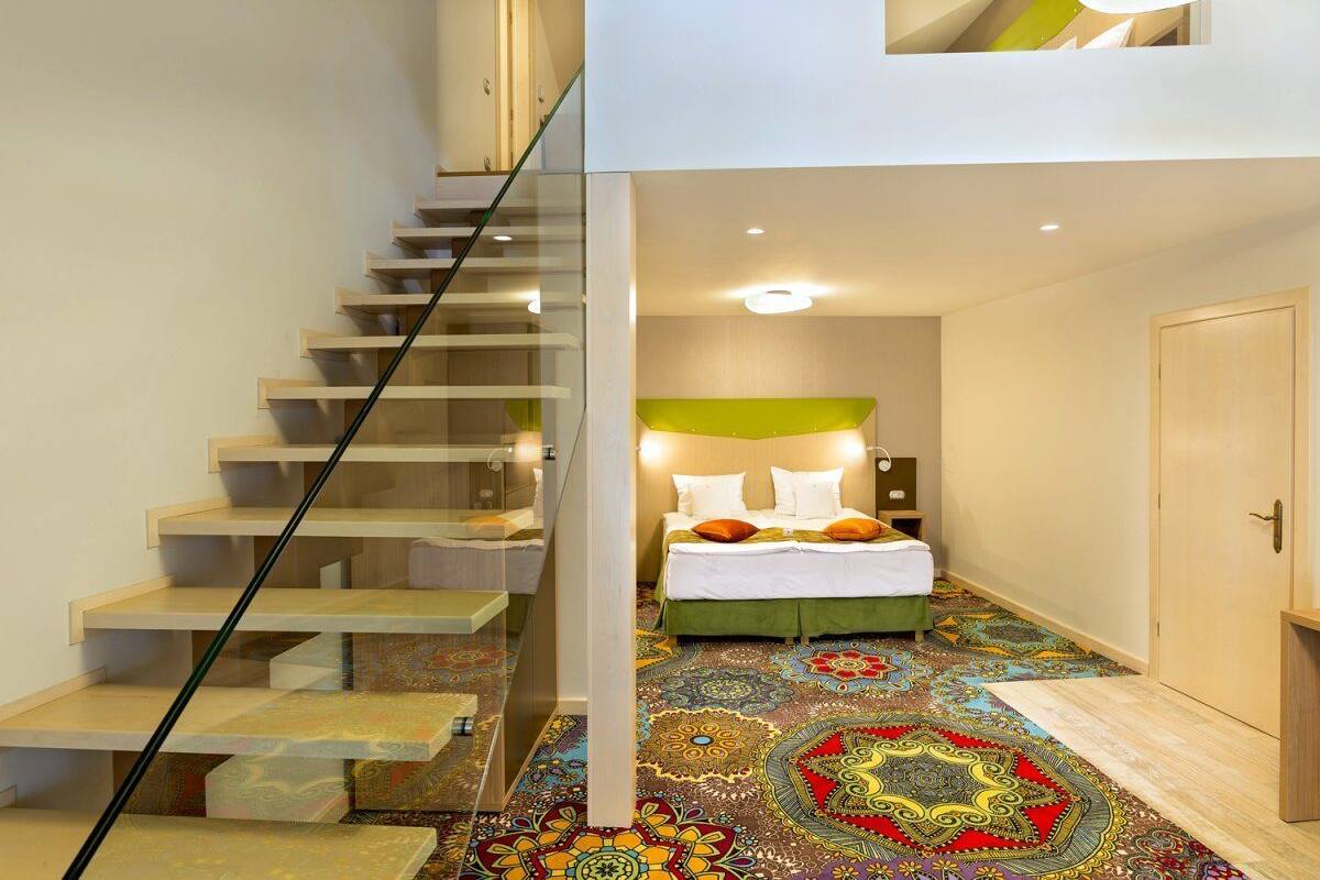 A modern hotel room with a staircase leading to a balcony, a bed with a green headboard, and a colorful patterned carpet.