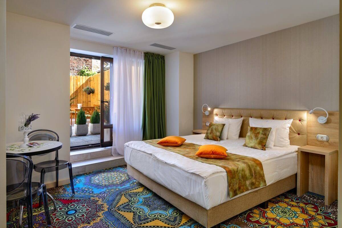 A hotel room with a large bed, a small table with chairs, a patterned carpet, and a view of the outside.
