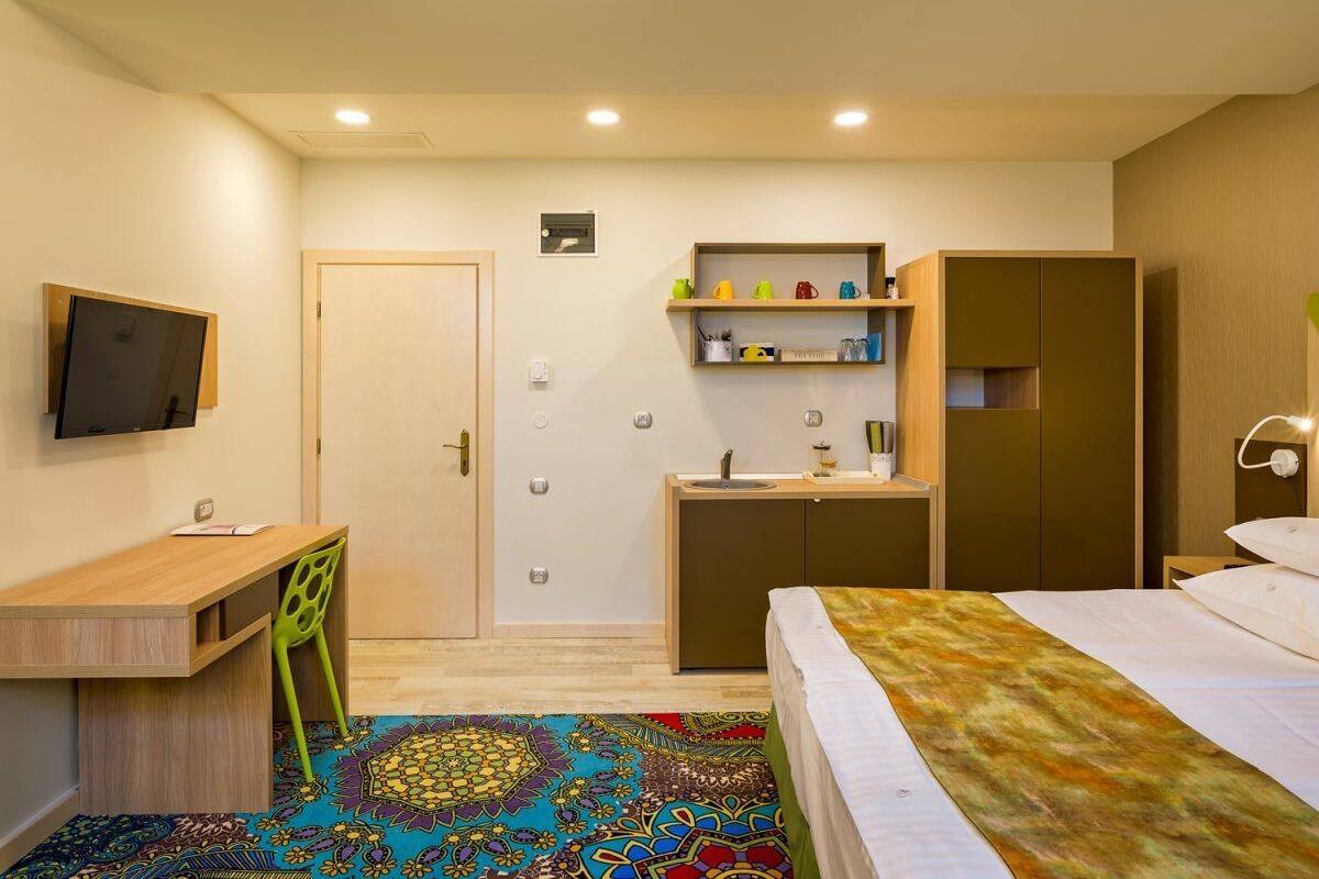 A hotel room with a bed, a desk with a chair, a TV mounted on the wall, a wardrobe, and a small kitchenette.