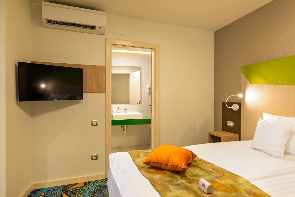 A hotel room with a bed, a TV mounted on the wall, a bedside table with a lamp, and a bathroom visible through an open door.