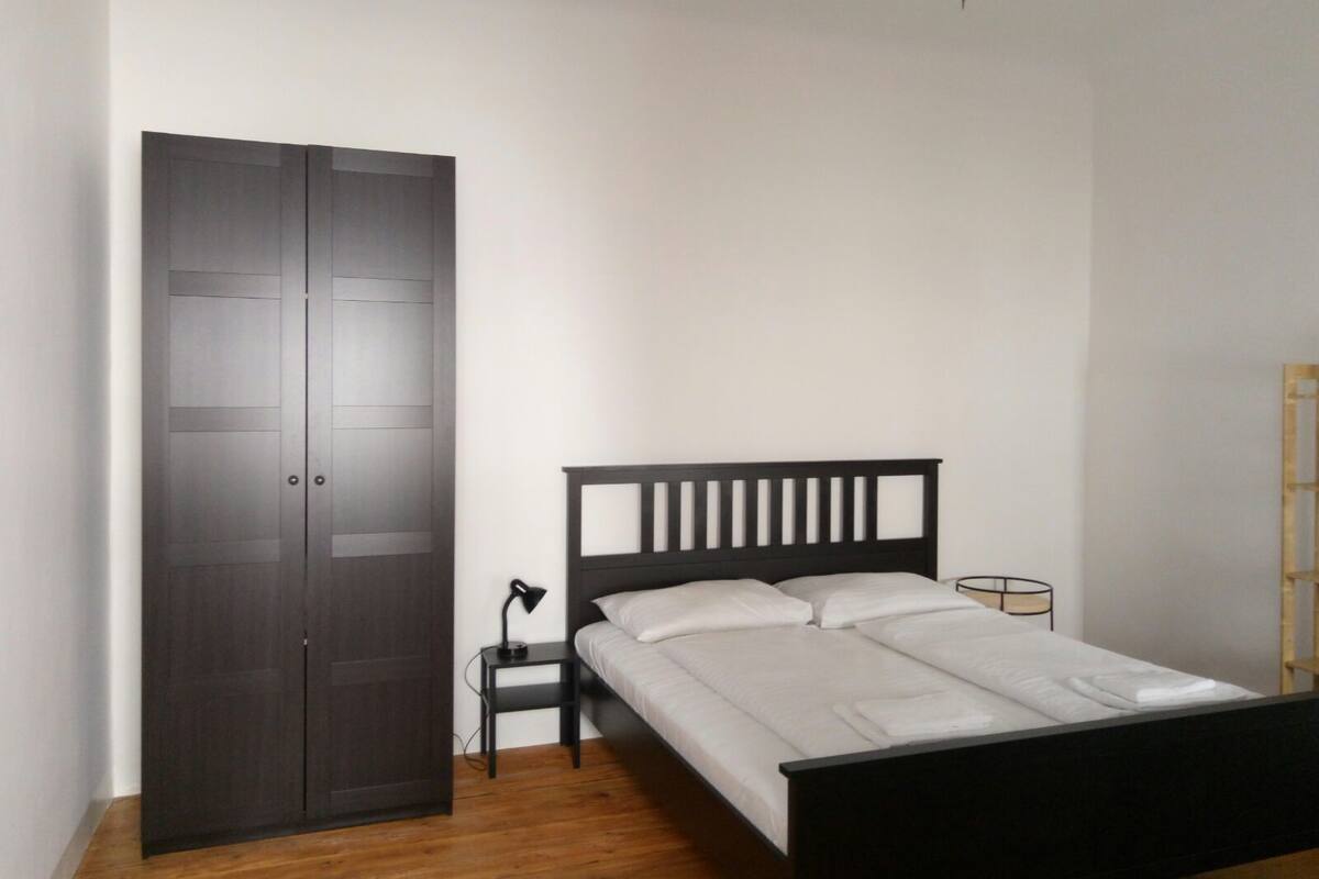 A bedroom with a bed, a wardrobe, a nightstand, a lamp, and a chandelier.