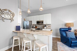 A modern kitchen with white cabinets, a marble countertop, and a built-in microwave. There are two white stools and a blue leather chair with a patterned pillow.