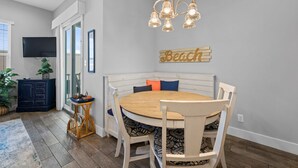 Un comedor con una mesa redonda de madera, sillas blancas tapizadas, un banco de madera con cojines, una lámpara de araña, un televisor de pantalla plana y un letrero en la pared que dice 'Beach'.