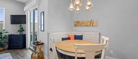 Un coin repas avec une table ronde en bois, des chaises à revêtement blanc, un banc en bois avec des coussins, un lustre, un téléviseur à écran plat et un panneau décoratif représentant une plage, accroché au mur.