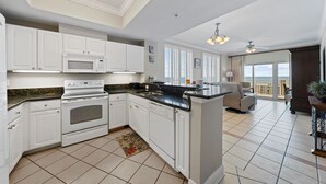 A modern kitchen with white cabinets, a built-in microwave, and a stove. There is a dining area with a view of the ocean. - Corner Condo with Panoramic Gulf & Lagoon Views | Sleeps 6 (Gulf Shores)