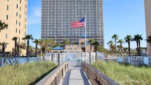 A hotel with a large American flag, a boardwalk leading to the entrance, and palm trees around. - Corner Condo with Panoramic Gulf & Lagoon Views | Sleeps 6 (Gulf Shores)
