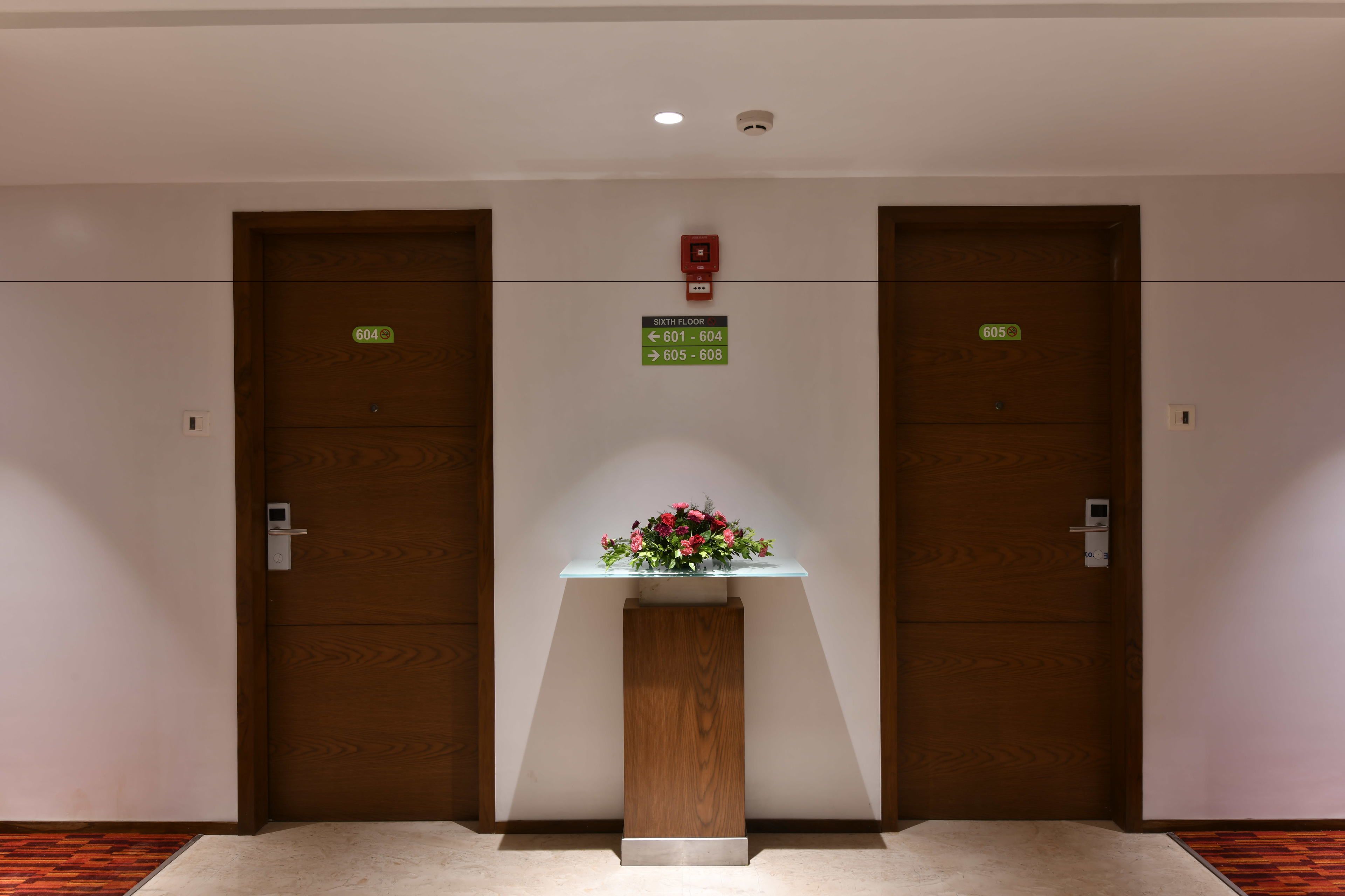 A hallway with two wooden doors, each marked with a number and an arrow indicating the direction to the sixth floor. A small pedestal with a vase of flowers is centered between the doors.