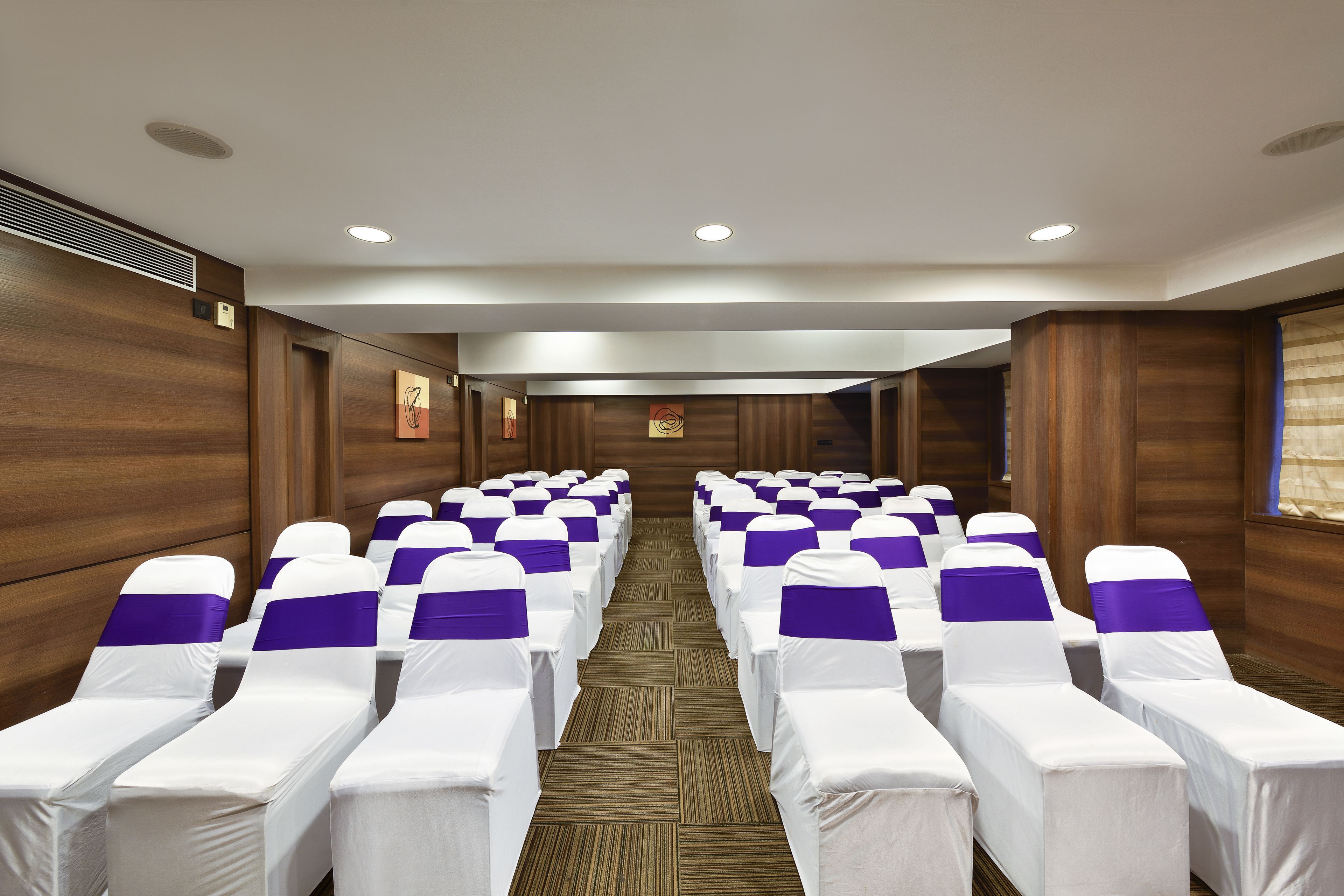 A hotel conference room with rows of white chairs with purple sashes, wooden panel walls, and recessed ceiling lights.