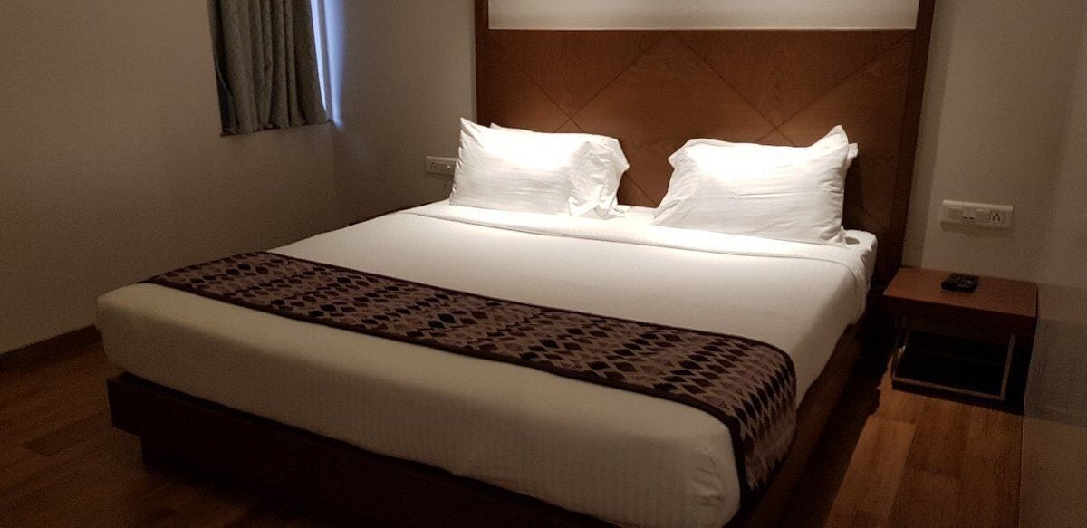 Un letto rifatto con lenzuola bianche e una coperta decorata, una testiera in legno, un comodino con un telecomando e una presa elettrica sulla parete.