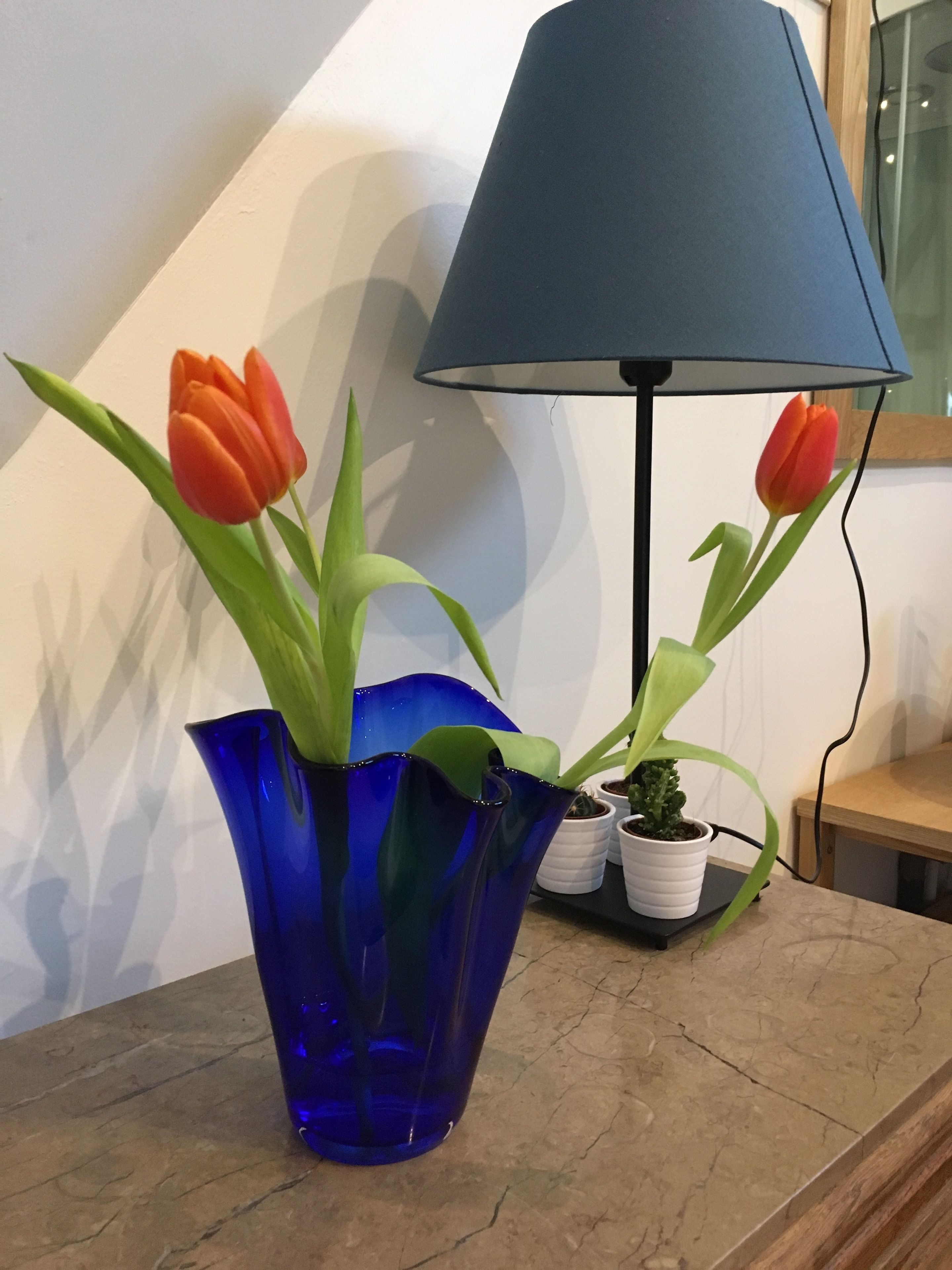 Un vase bleu contenant des tulipes orange, posé sur une table en bois, avec une lampe et de petites plantes en pot.