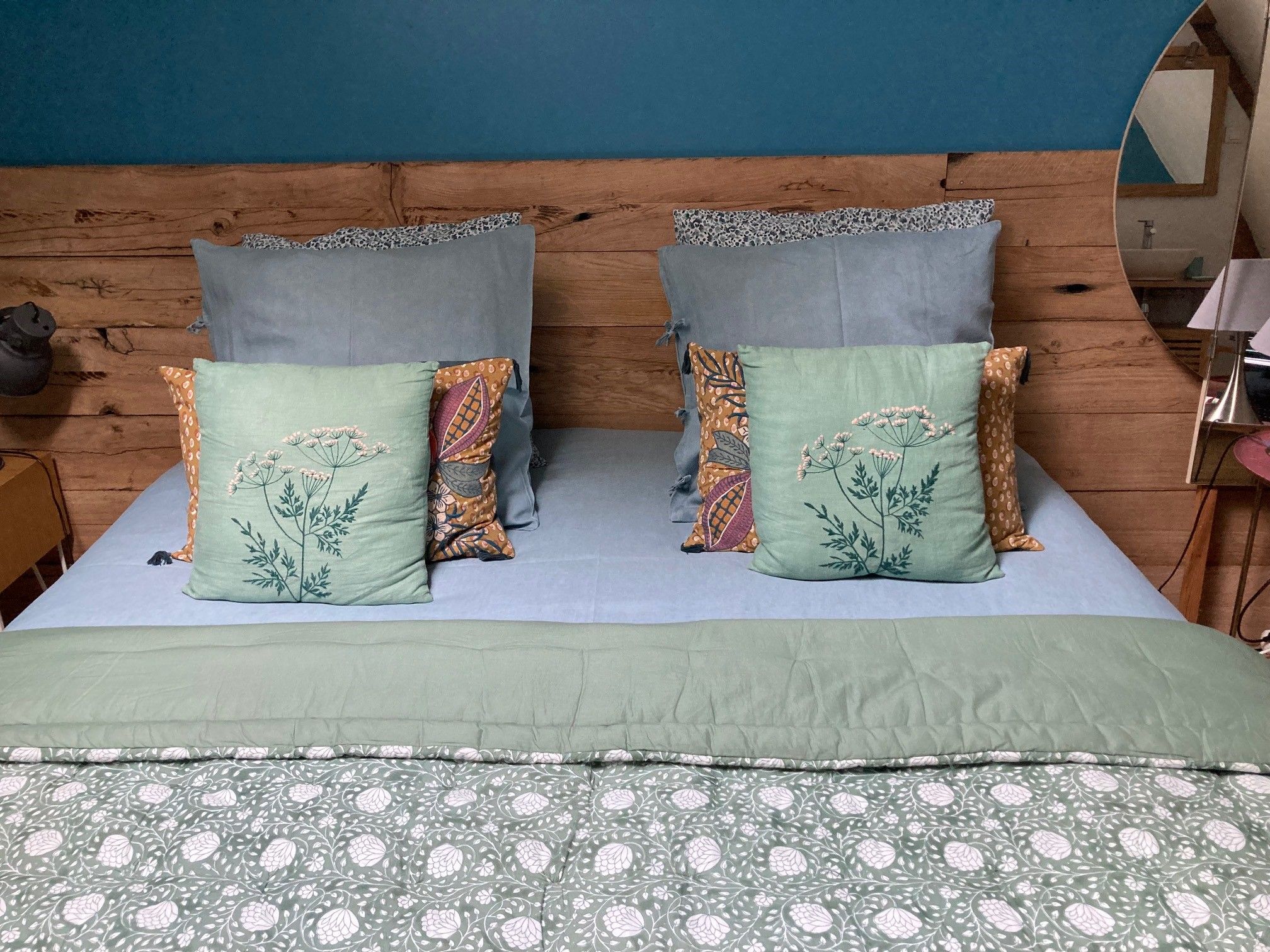 Een bed met een blauw hoofdeinde, twee kussens met bloemenborduurwerk en een groene sprei met een bloemendesign.