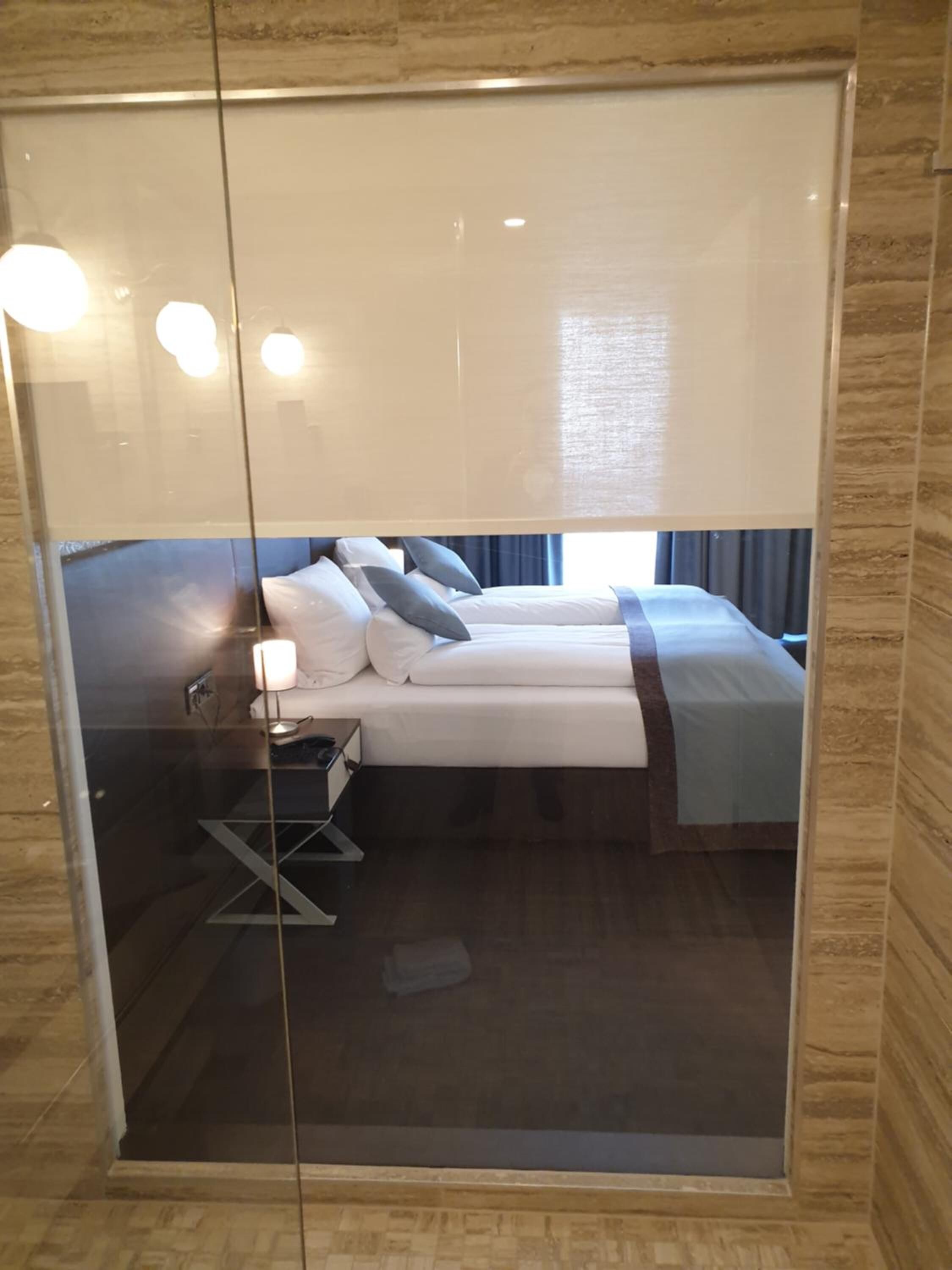 A hotel room with a large mirror, a bed with white linens, a bedside table with a lamp, and a view of the outside through the window.