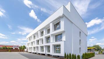 Un edificio moderno y blanco de varios pisos con amplias ventanas, rodeado de un área pavimentada y vegetación.