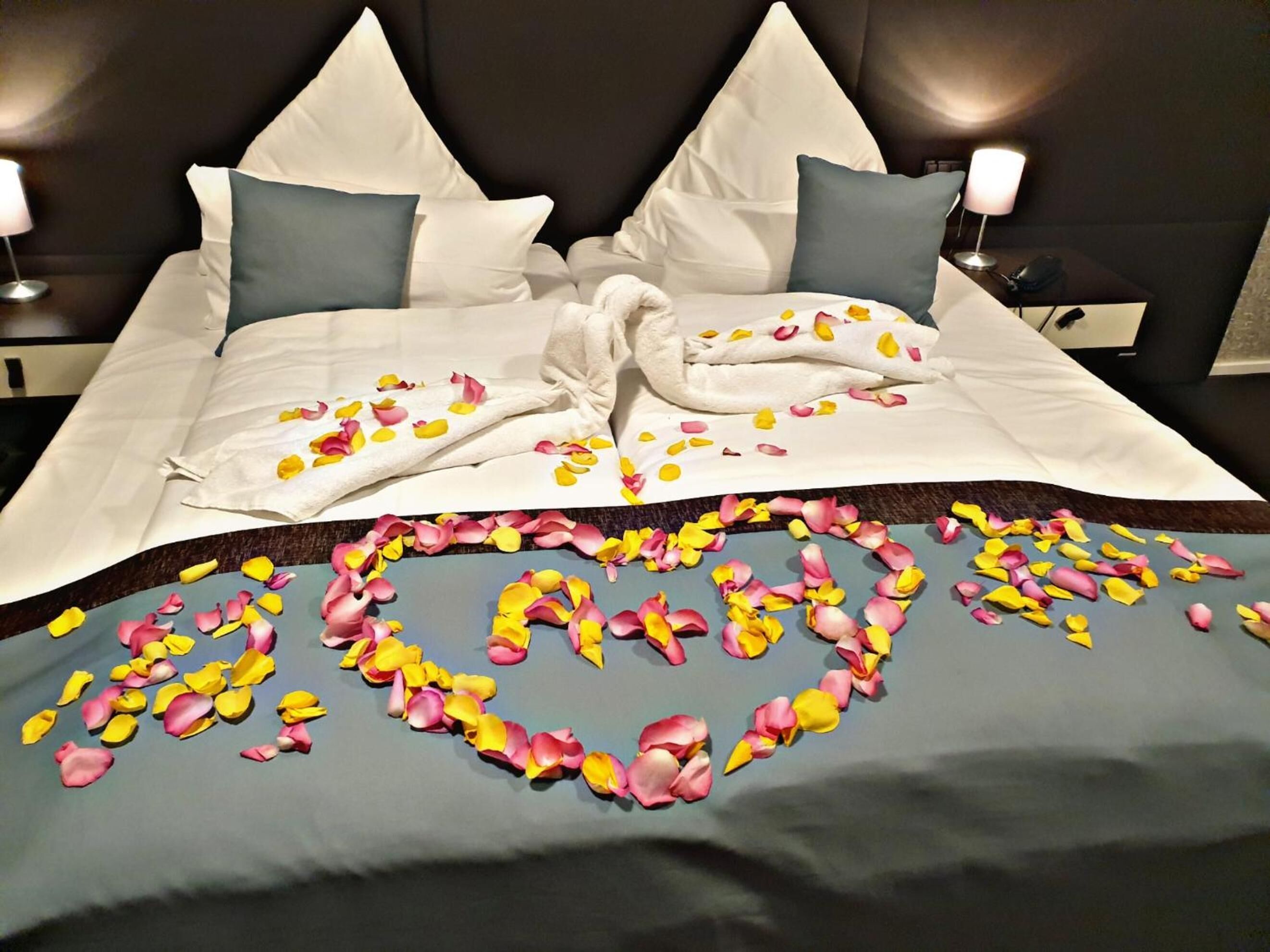 Un letto rifatto con cura, lenzuola bianche, due cuscini e un composizione a forma di cuore di petali di rosa sul coprimaterasso.