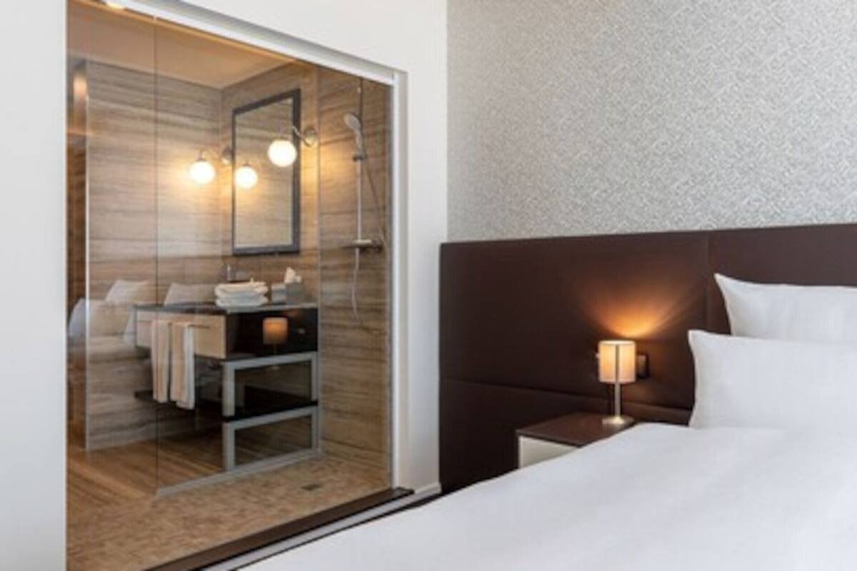Habitación de hotel con una cama grande, una mesita de noche con lámpara, cabecero de madera y una mampara de ducha de vidrio con espejo e iluminación.