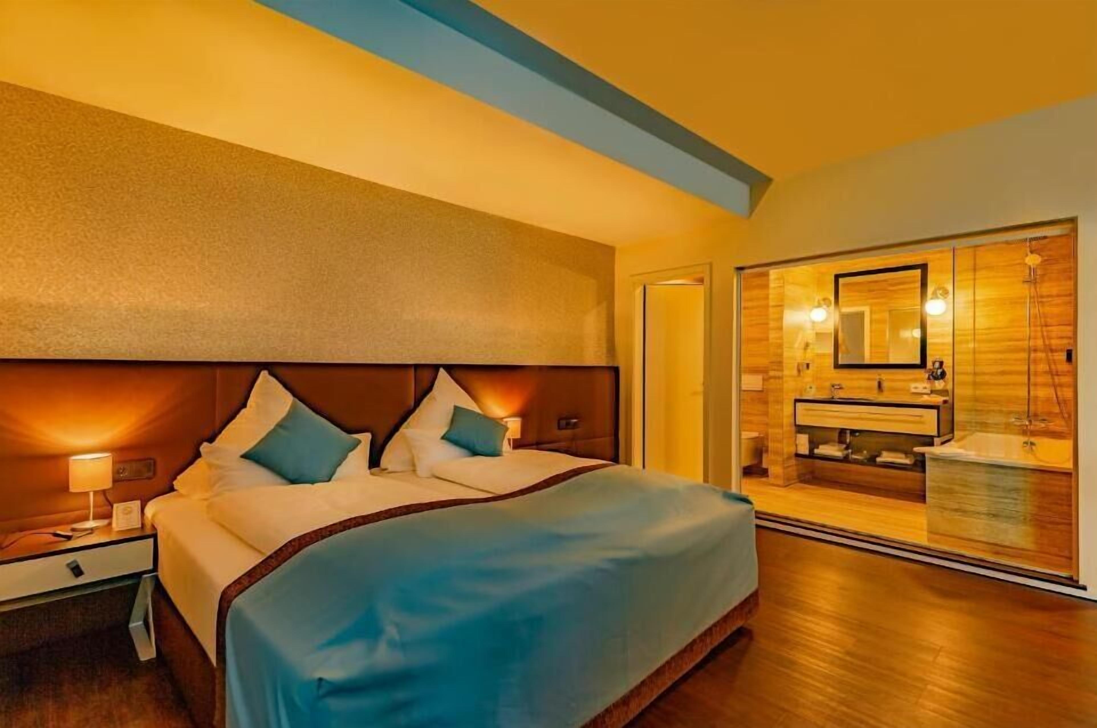 Habitación de hotel con una cama grande, una mesita de noche con lámpara y un baño con espejo y lavamanos.