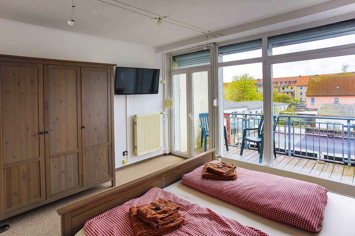 Une chambre avec deux lits, une armoire en bois, une télévision et un balcon équipé de chaises et d’une rambarde.