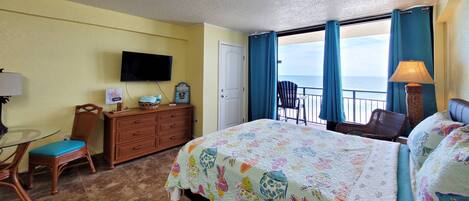 A hotel room with a bed, a television, a desk with a chair, a dresser, and a view of the ocean through large windows.