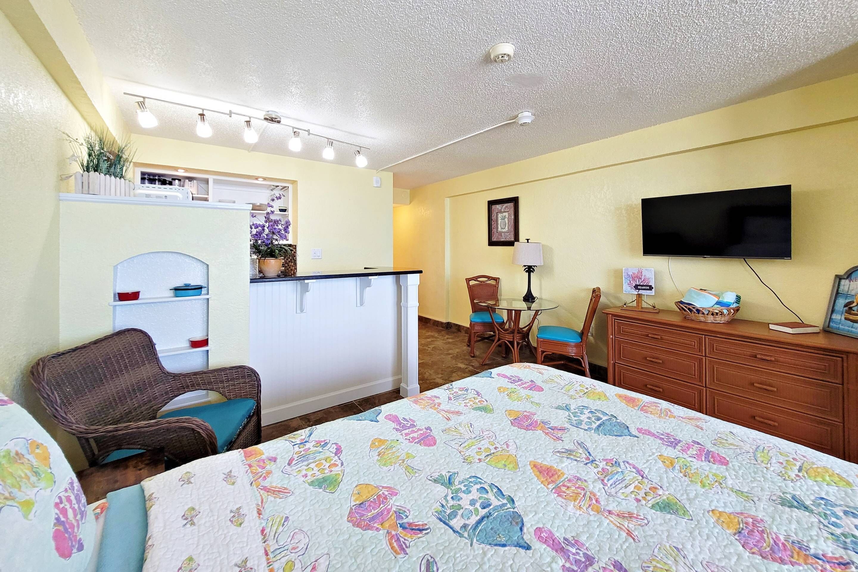 Habitación de hotel con cama, una silla, escritorio con lámpara, televisor y kitchenette.