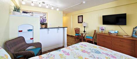 Habitación de hotel con cama, una silla, escritorio con lámpara, televisor y kitchenette.