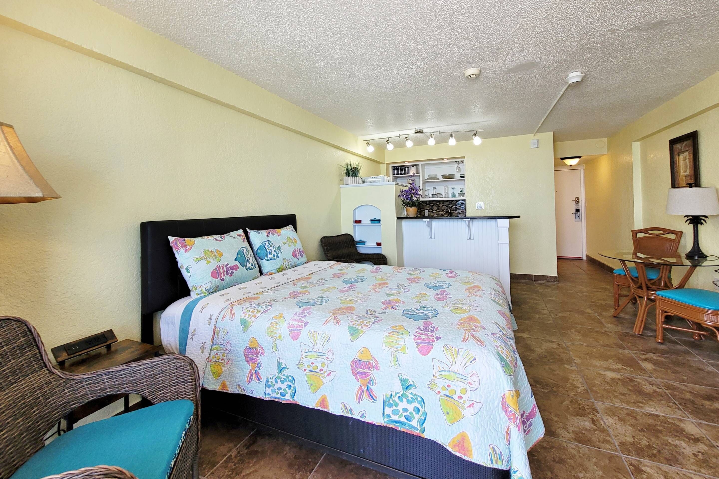 A bedroom with a bed, a chair, a small table, and a kitchenette in the background.