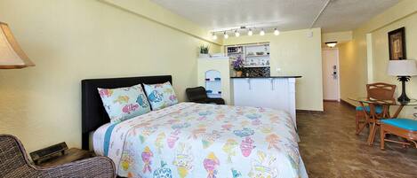 A bedroom with a bed, a chair, a small table, and a kitchenette in the background.
