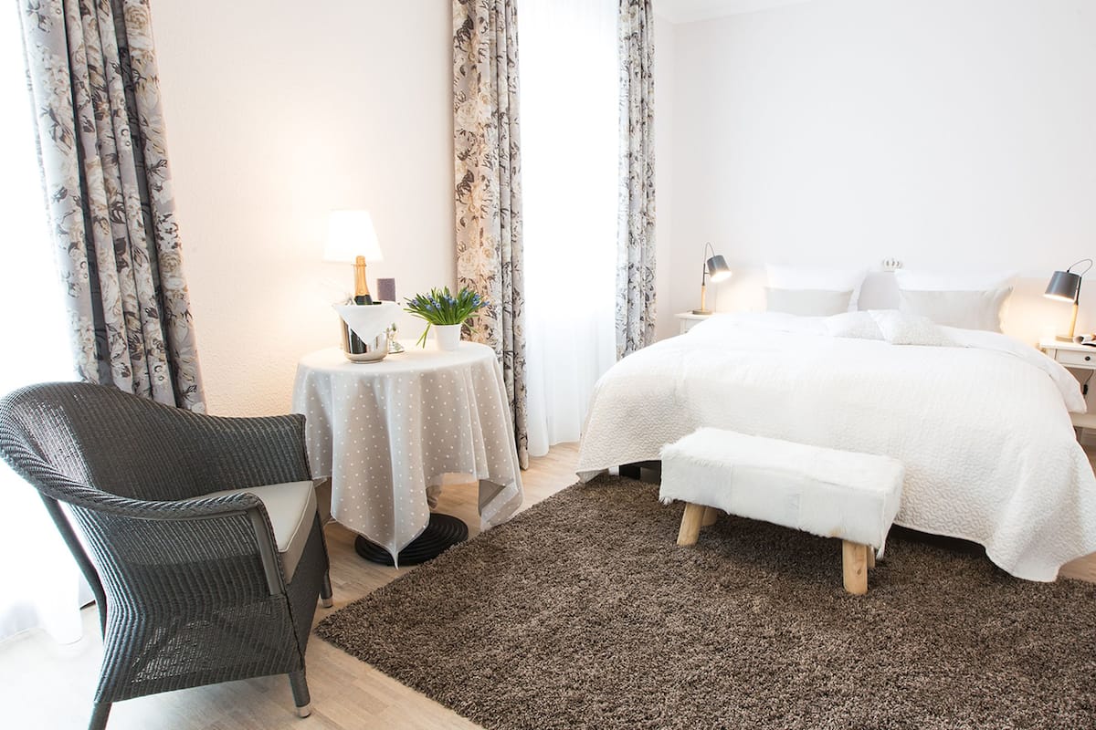 A bedroom with a large bed, a bedside table with a lamp, a small round table with a lamp, a chair, and a rug.