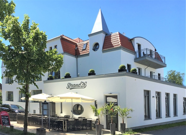 A white building with a red-tiled roof, a balcony, and outdoor seating with umbrellas.