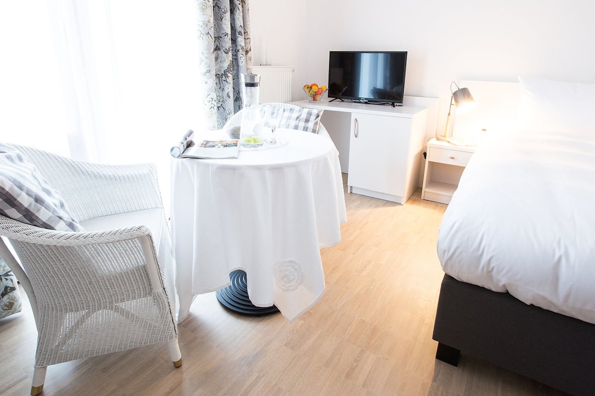 A hotel room with a bed, a round table with a white tablecloth, a wicker chair, a television on a cabinet, and a nightstand with a lamp.