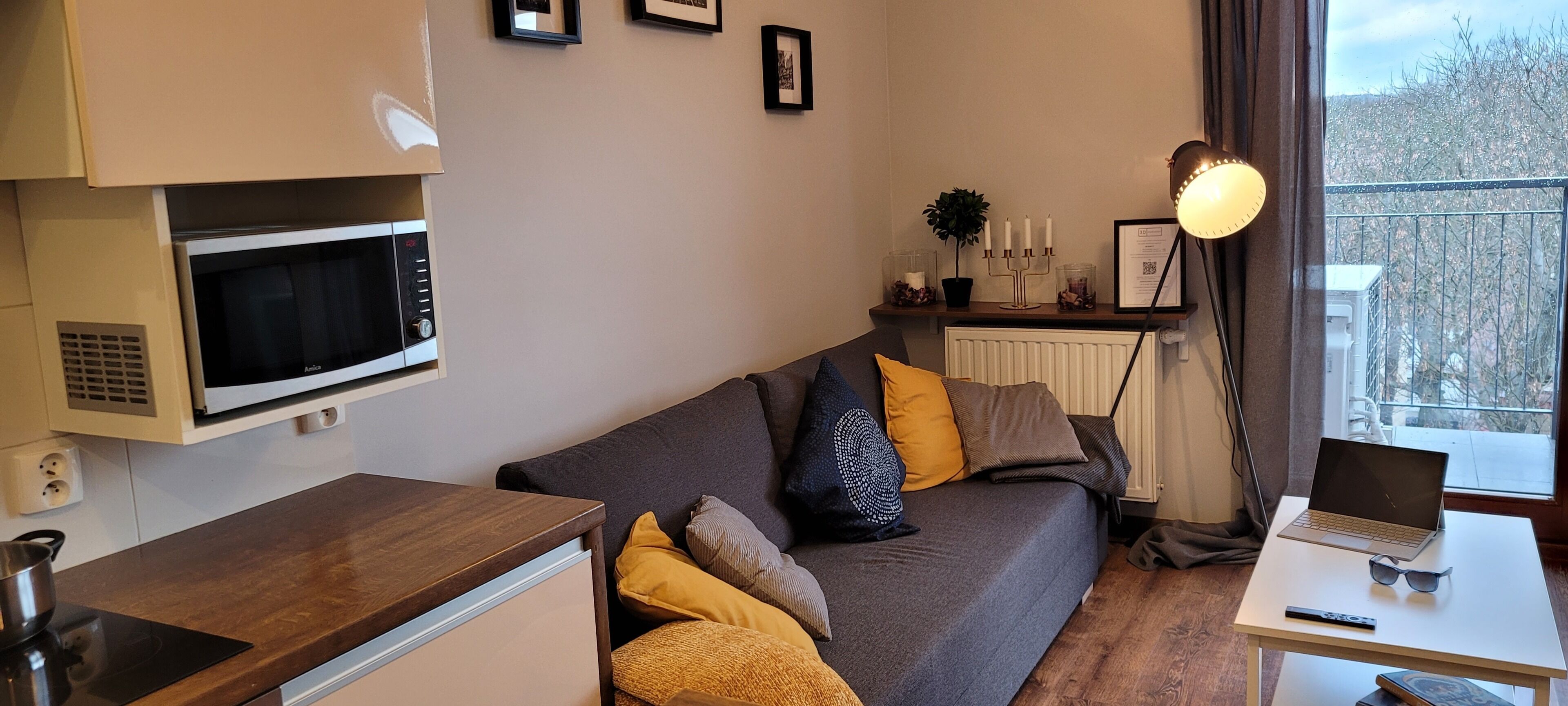 A compact living space with a kitchenette, a sofa with cushions, a dining table with a laptop and sunglasses, and a view of the outside.