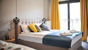 A hotel room with a large bed, a bedside table with a lamp, and a view of the outside through a window with yellow curtains. - 3D Szlak Apartament (Kraków)