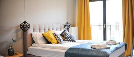 Een hotelkamer met een groot bed, een nachtkastje met een lamp, en uitzicht naar buiten door een raam met gele gordijnen.