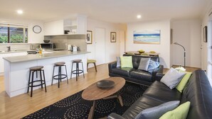 Un apartament modern amb cuina, zona de dinar i saló. Inclou un sofà de pell negra, una taula de centre de fusta i una illa de cuina amb tamborets.