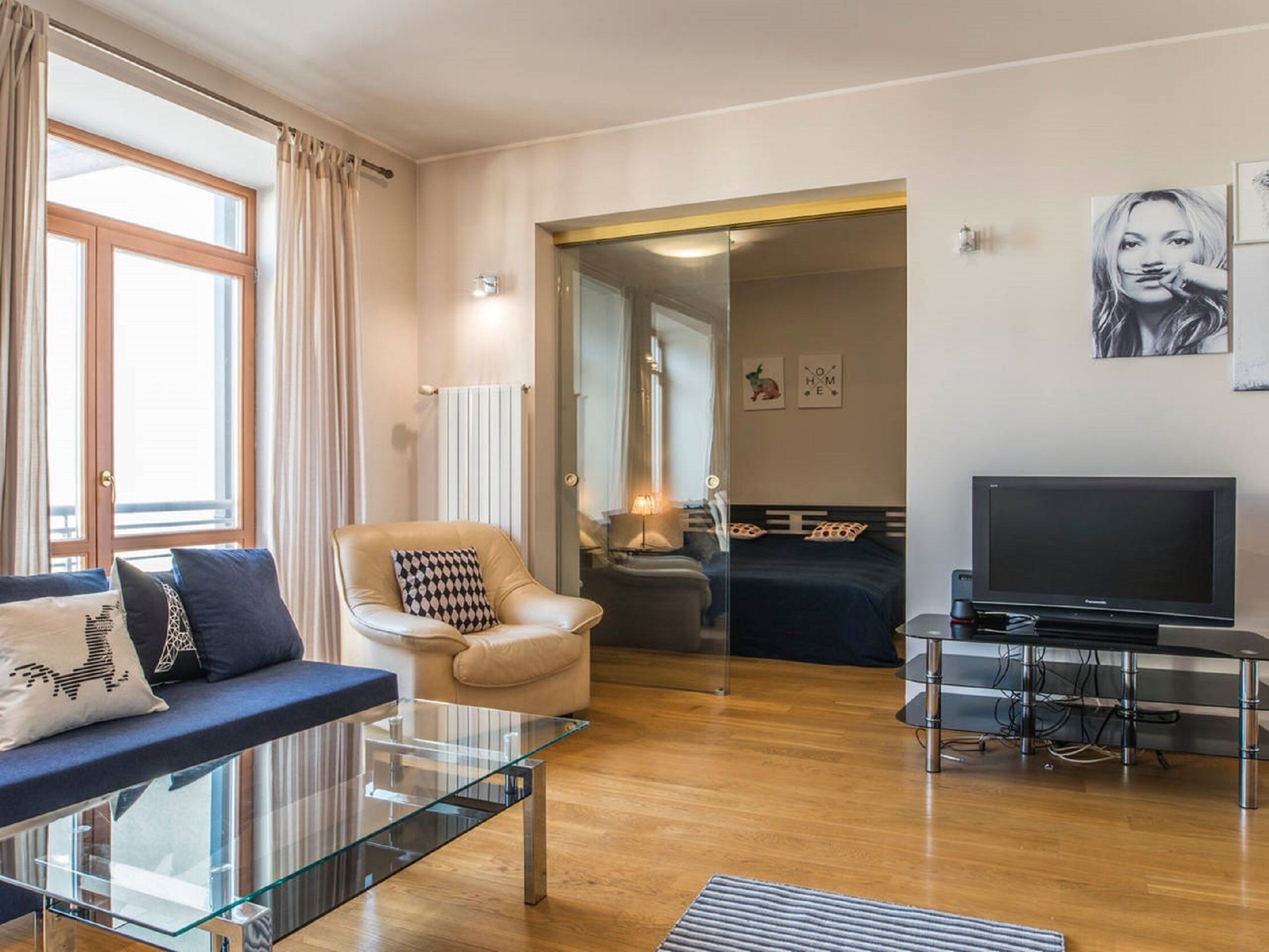 Un salon moderne avec une table basse en verre, un fauteuil en cuir, un canapé agrémenté de coussins à motifs, un téléviseur à écran plat et un lit recouvert d’une couvre-lit bleu foncé.