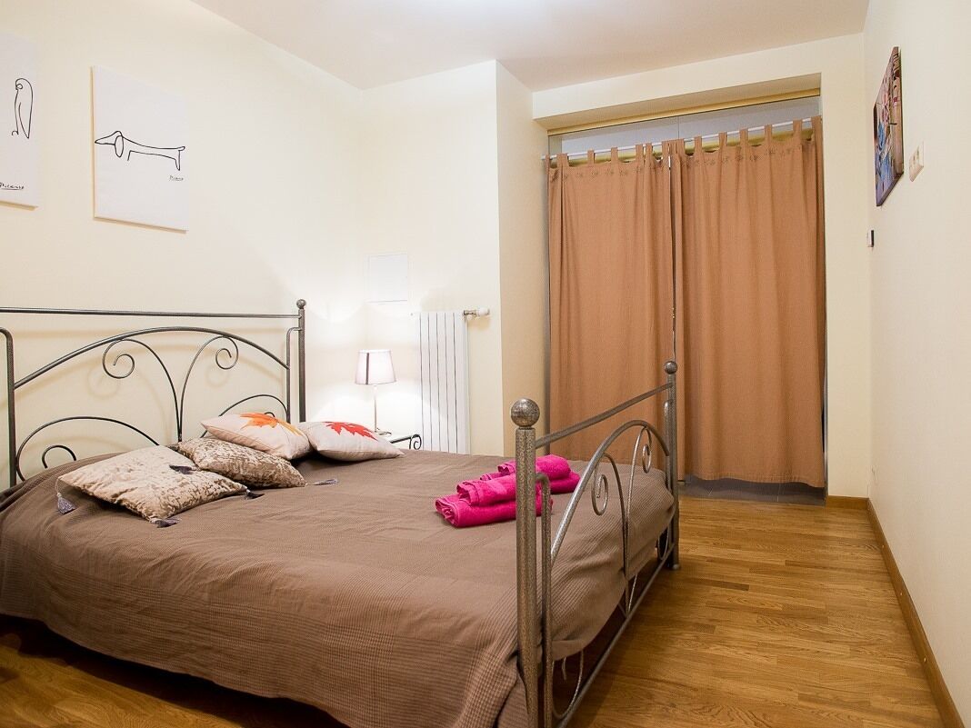 A bedroom with a metal bed frame, a brown bedspread, and pillows. There is a window with brown curtains, a radiator, and a wall-mounted lamp.