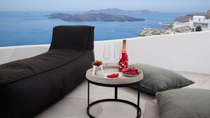 A balcony with a black sofa, a small round table with a bottle, glasses, and fruit. The view includes the sea and distant islands. - Amity Suites - Adults Only (Santorini)