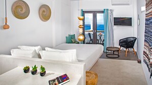 A modern hotel room with a bed, a small seating area with a table, and a view of the sea through a glass door. - Amity Suites - Adults Only (Santorini)