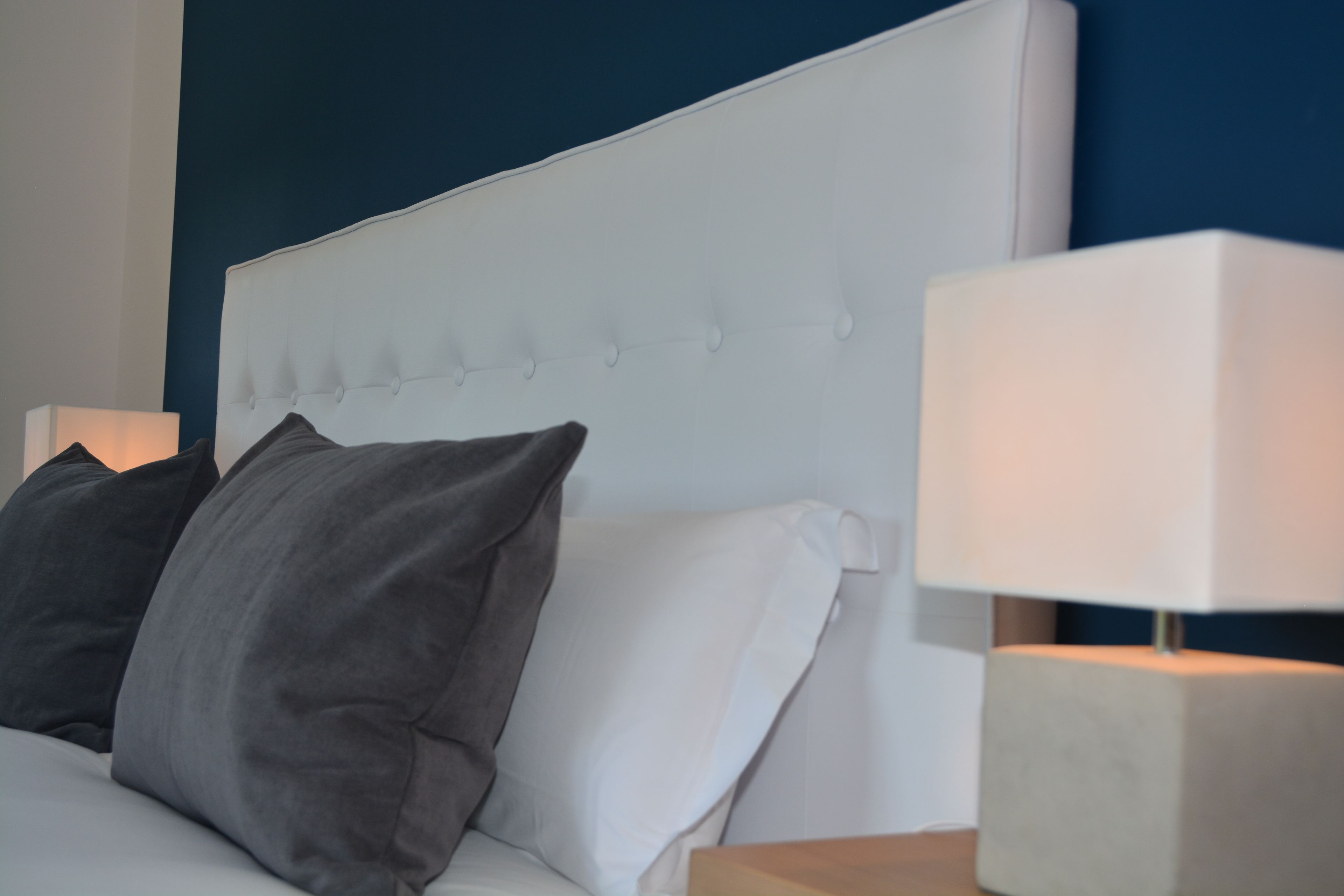 Een bed met een gestoffeerd hoofdbord, twee kussens en een nachtkastje met een lamp.