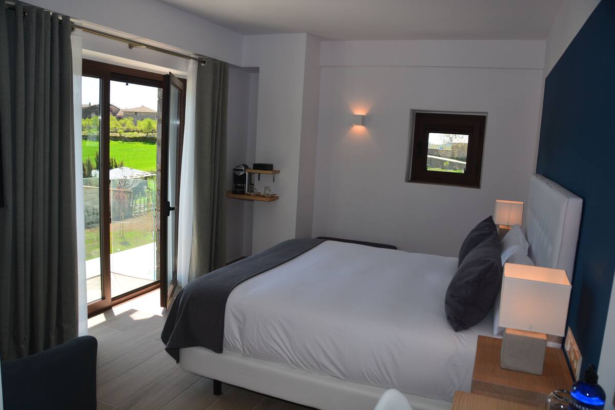 Een hotelkamer met een bed, een nachtkastje met een lamp, een klein plankje met spullen, en uitzicht op groen.