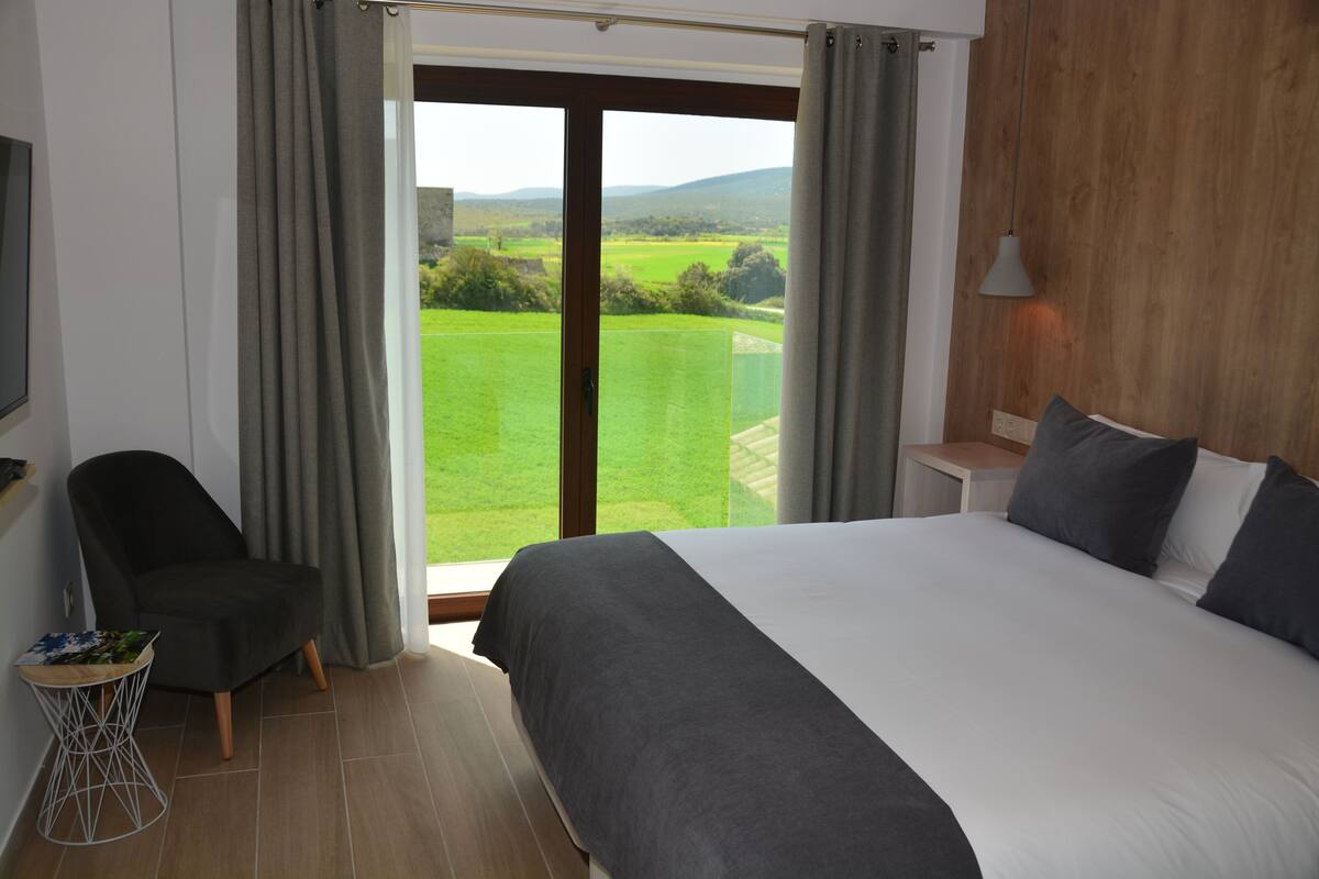 Slaapkamer met een groot bed, een stoel, een klein tafeltje en uitzicht op een groen landschap door een schuifdeur.