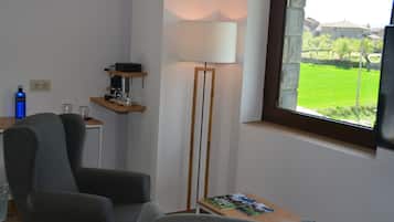 Una habitación con un sillón gris, una lámpara de pie, una mesita con una revista y una ventana con vistas a zonas verdes y edificios.