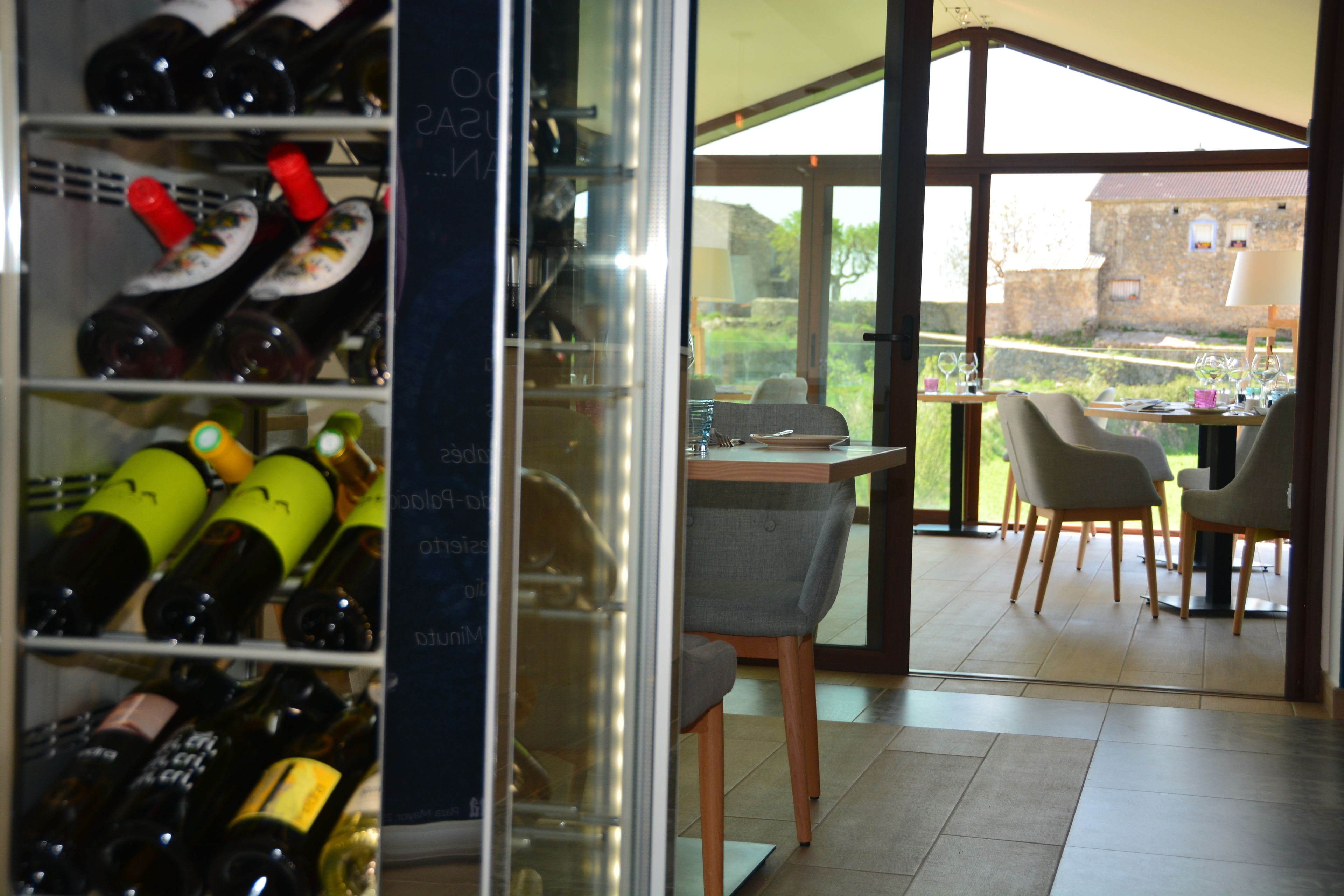 Une cave à vin avec des étagères remplies de bouteilles, un coin repas avec une table et des chaises, et une vue sur un bâtiment en pierre à l’extérieur.