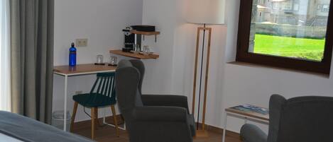 A hotel room with a bed, two armchairs, a desk with a coffee machine, a lamp, and a window with a view of buildings and a green lawn.