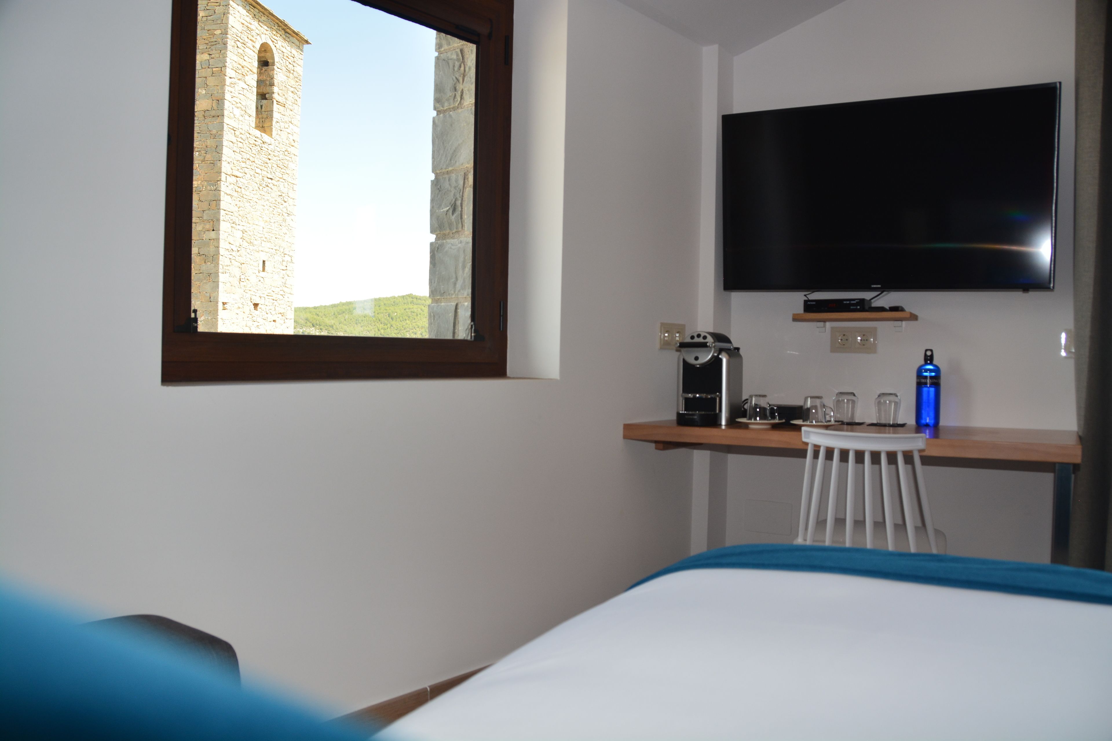 Habitación de hotel con cama, escritorio con cafetera, televisor montado en la pared y ventana con vista a una torre de piedra.