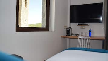 Habitación de hotel con cama, escritorio con cafetera, televisor montado en la pared y ventana con vista a una torre de piedra.