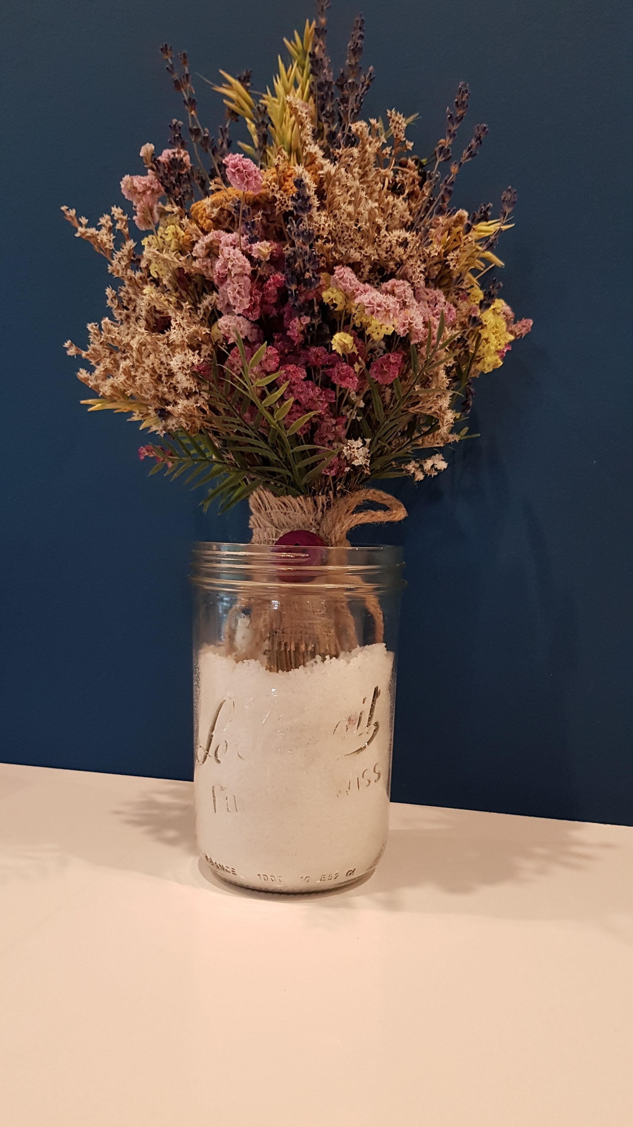 A mason jar filled with dried flowers and greenery, placed on a table against a blue wall.