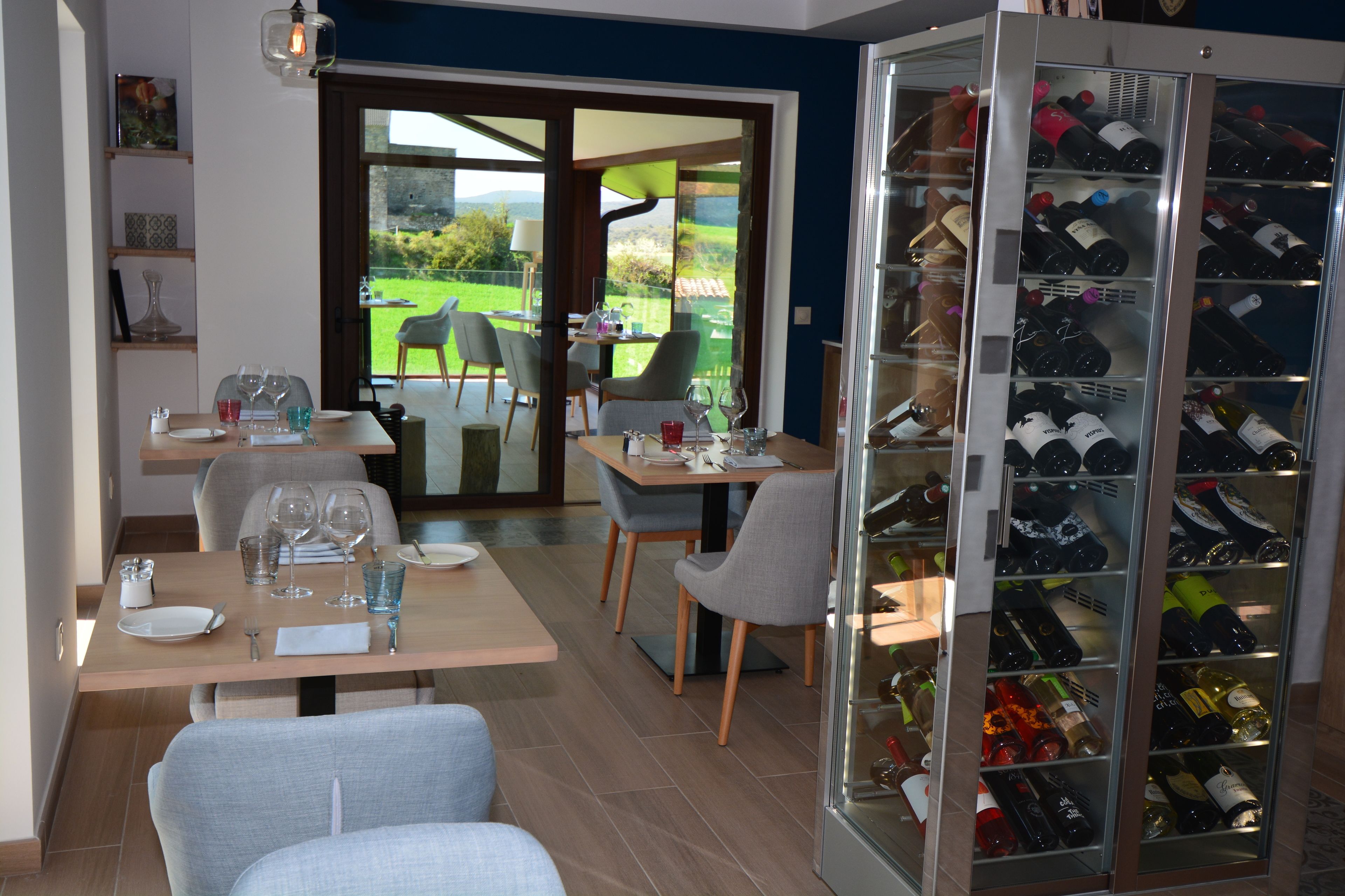 A dining area with a table set for two, a wine refrigerator stocked with bottles, and a view of a green landscape through the glass door.
