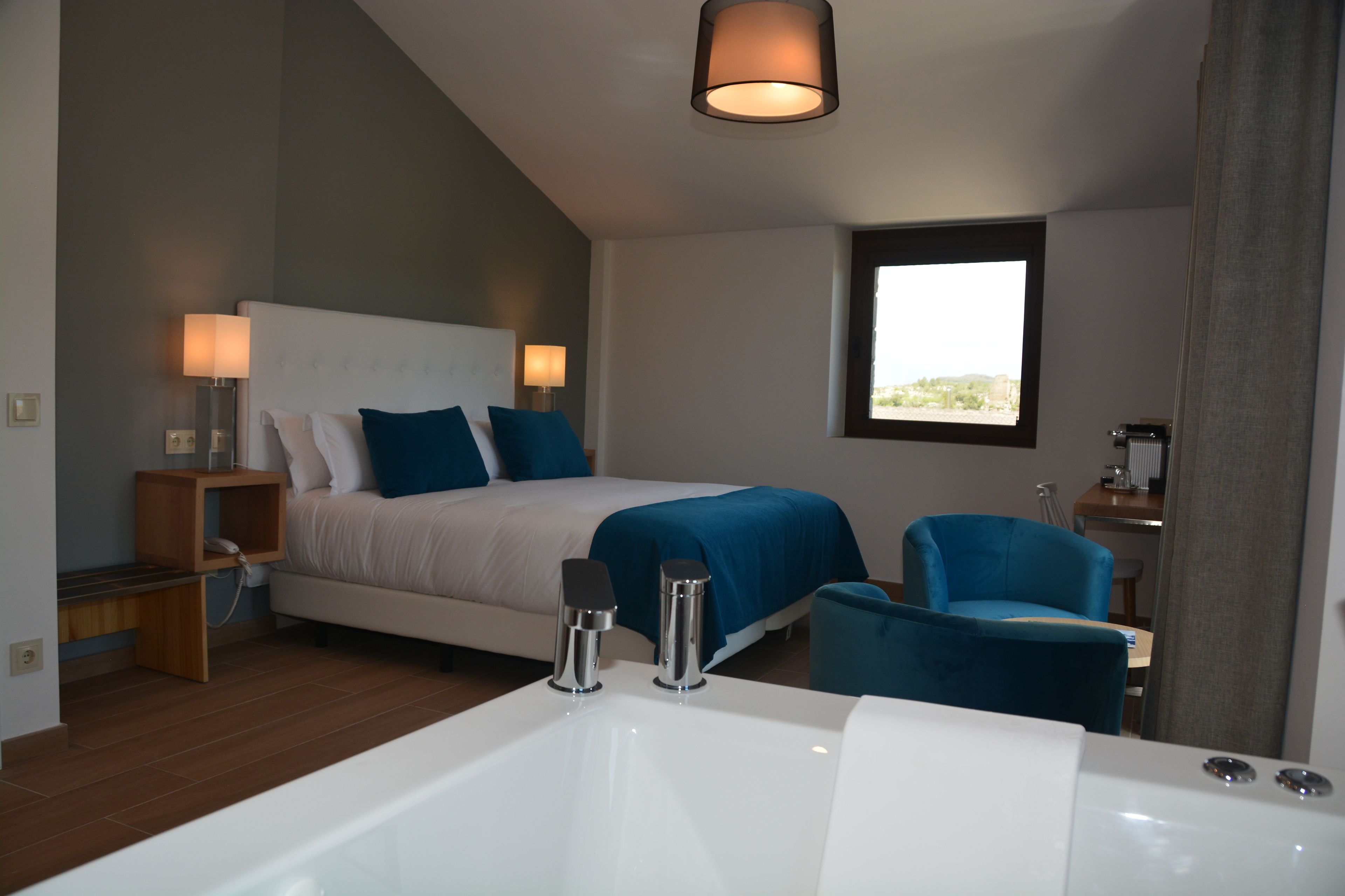 Habitació d'hotel moderna amb un llit gran, zona de seients amb una cadira blava, una banyera espaiosa i una finestra amb vista.