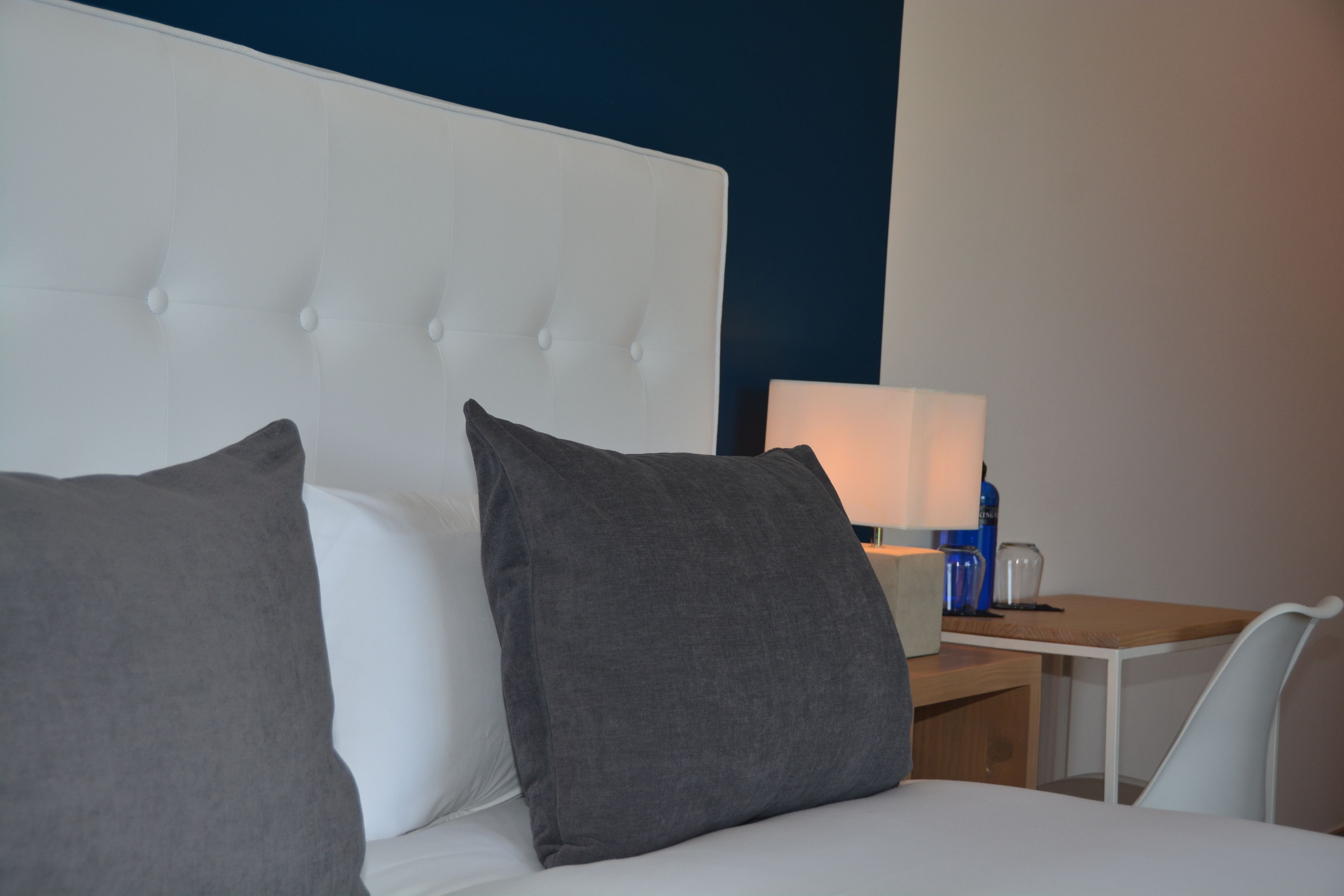 Habitació d'hotel amb un llit blanc, almoinades grises, una taula de nit amb una làmpada i una cadira.