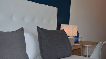 Habitació d'hotel amb un llit blanc, almoinades grises, una taula de nit amb una làmpada i una cadira.